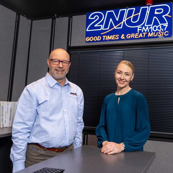 Ian Crouch and Cecilia Connell smiling toward the camera in the 2NURFM radio station. There is a blue and yellow neon sign above them that reads '2NURFM103.7 Good Times & Great Music.'. 30-year legacy nurtures new broadcasting talent.