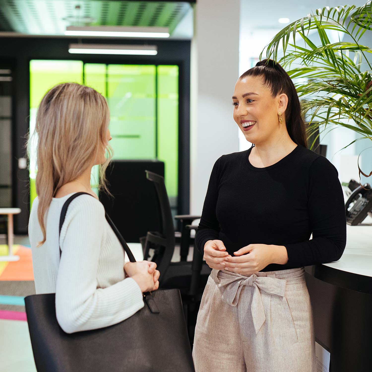 Two women standing and talking in a modern office space, one holding a black tote bag and the other smiling with hands clasped.