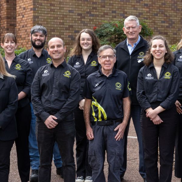 A group of twelve people stands outdoors in front of one of the buildings in the Engineering precinct, all wearing matching black shirts with a yellow and white Science and Engineering Challenge logo on the right and a University of Newcastle logo on the left. They pose together on a paved area with greenery behind them, appearing relaxed and friendly.