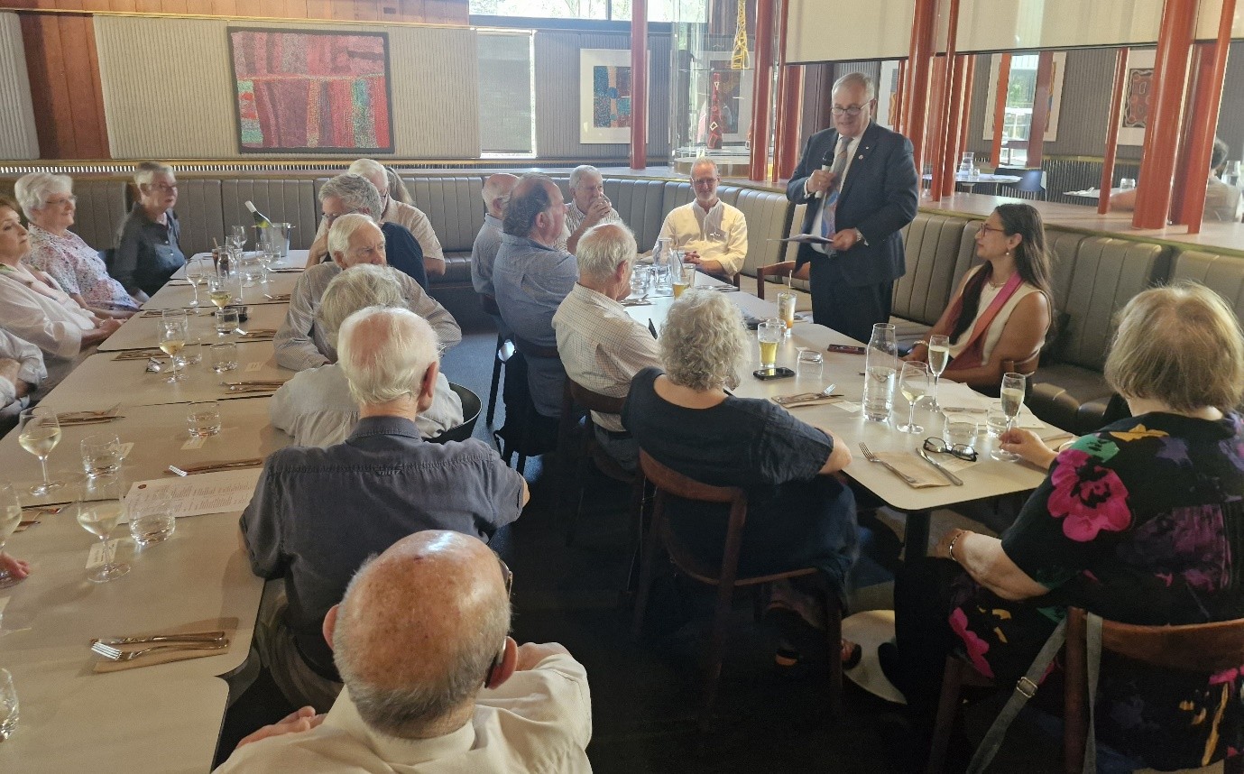 A group of NERLA members sit around a long table in a restaurant listening to the Vice-Chancellor who is standing and speaking