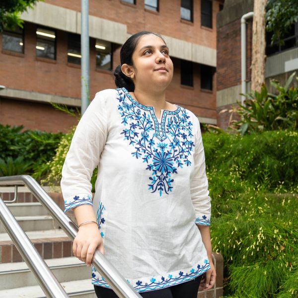 A person wearing a white tunic with blue embroidery stands on outdoor steps beside greenery and a brick university building.. From Delhi to Newcastle, full tuition scholarship makes STEM dreams come true.