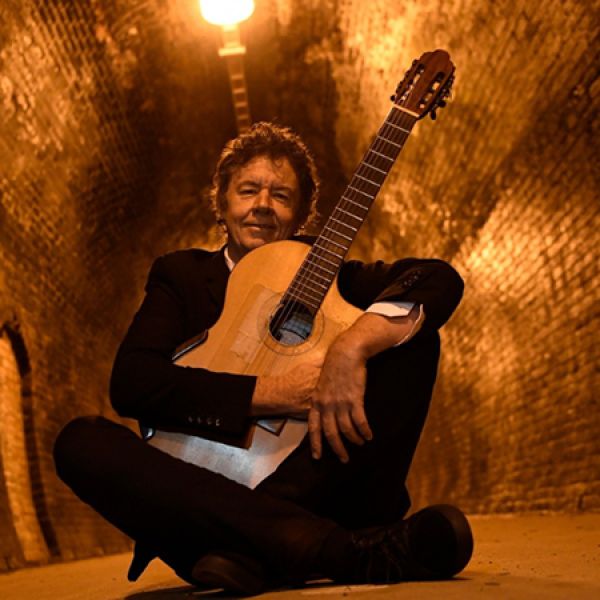 A person in a suit sits cross‑legged in a warmly lit tunnel while holding an acoustic guitar upright.