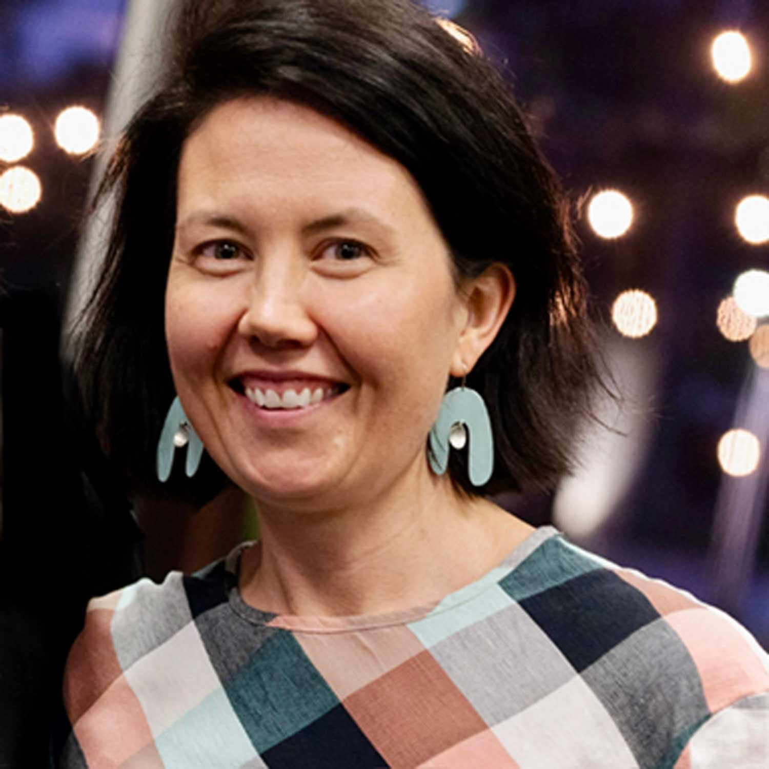 Brunette female smiling at the camera, wearing decorative arch shaped earrings and a multicoloured gingham pattern top. 