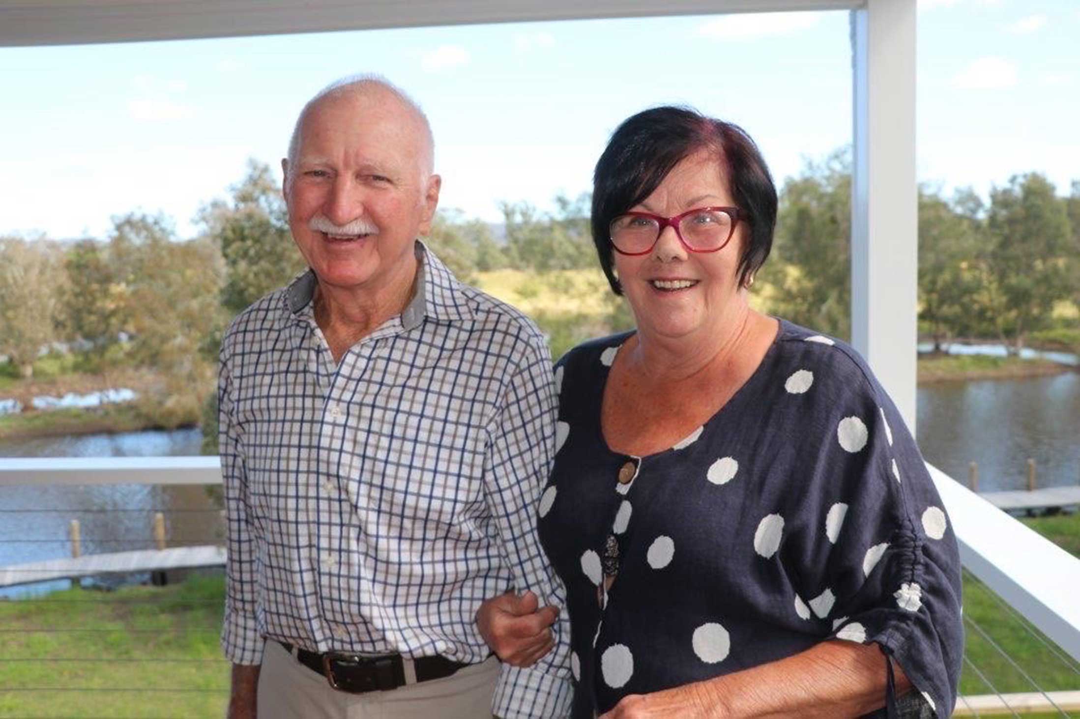 An older couple smiling and looking at the camera, standing on a balcony overlooking a lake. 