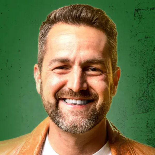Close-up studio portrait of a person with short brown hair and a neatly trimmed beard, smiling broadly against a textured green background while wearing a light shirt and tan jacket.