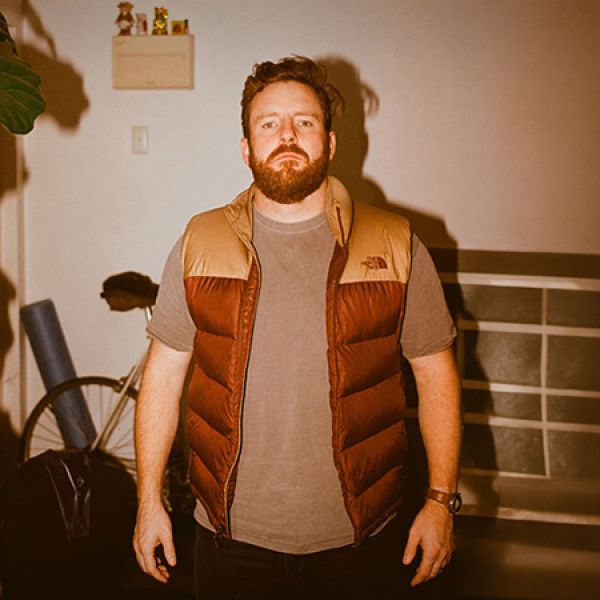 Indoor photo of a bearded person standing upright in a warmly lit room, wearing a brown puffer vest over a grey shirt, with a bicycle, plant, and shelving visible in the background.