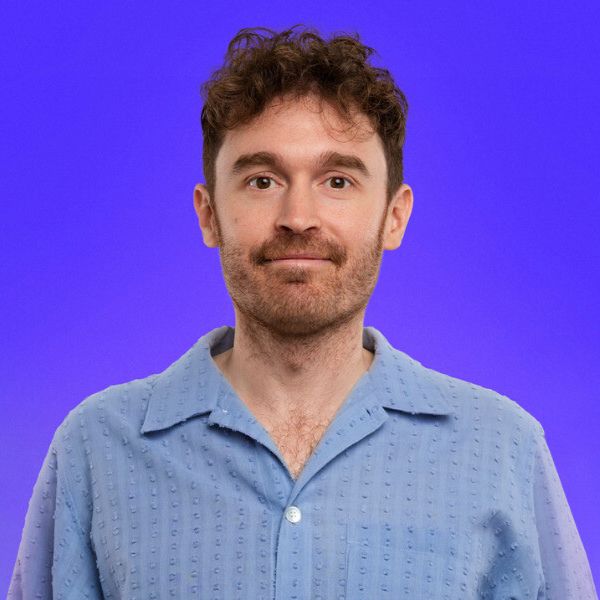 Studio portrait of a person with short curly hair and light facial hair, wearing a textured blue button-up shirt against a smooth purple background.