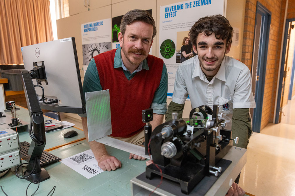 A high school student stands with a male Academic staff member, near equipment emitting a green laser