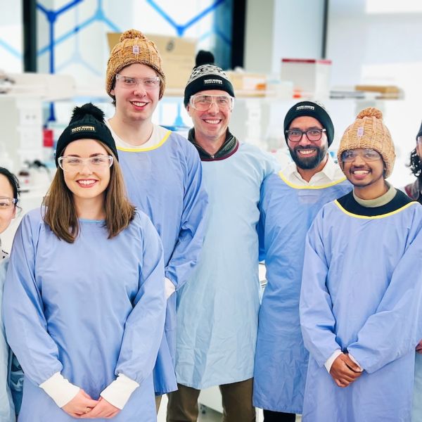 A group of people in a lab, wearing lab gowns and a mix of Mark Hughes Foundation beanies and caps. All are looking directly to camera and smiling