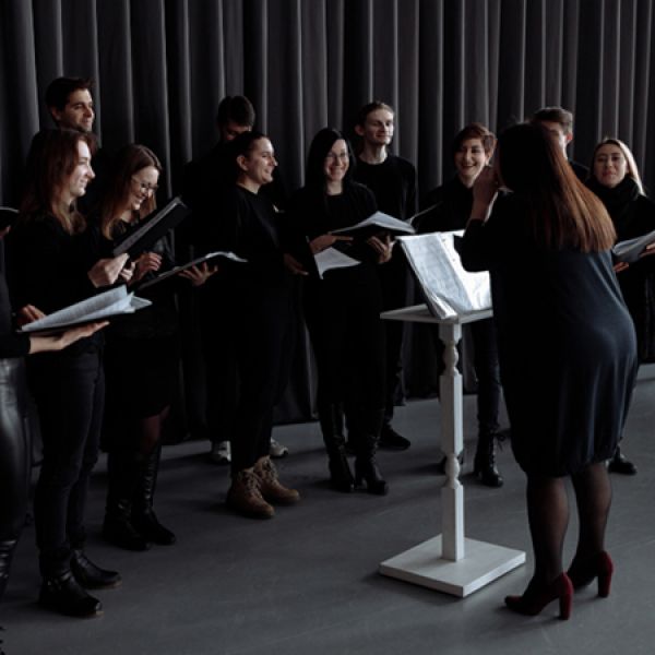 A choir stands in a semicircle holding sheet music while a conductor leads from a music stand in front of a dark curtained backdrop.