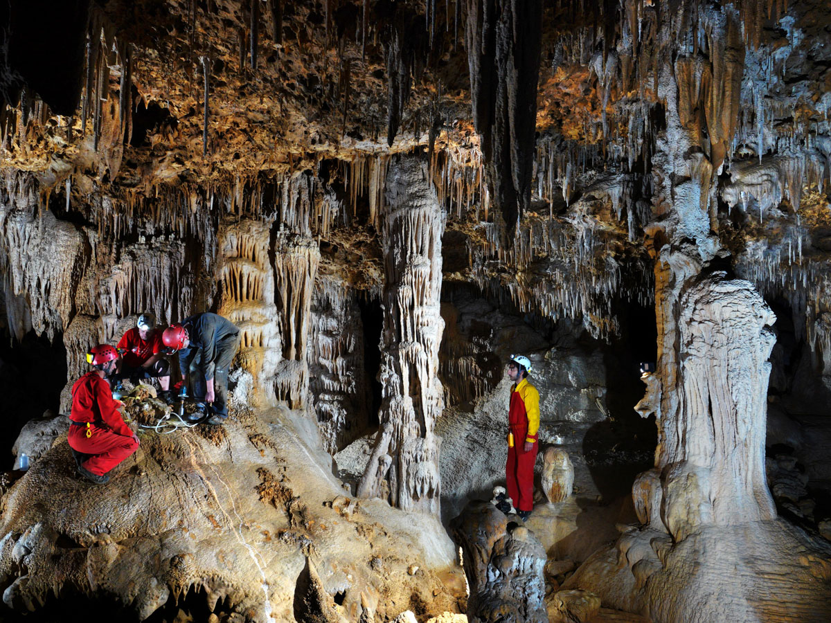 Four helmeted researchers explore a limestone cave filled with towering stalagmites, columns and delicate stalactites hanging from the ceiling.
