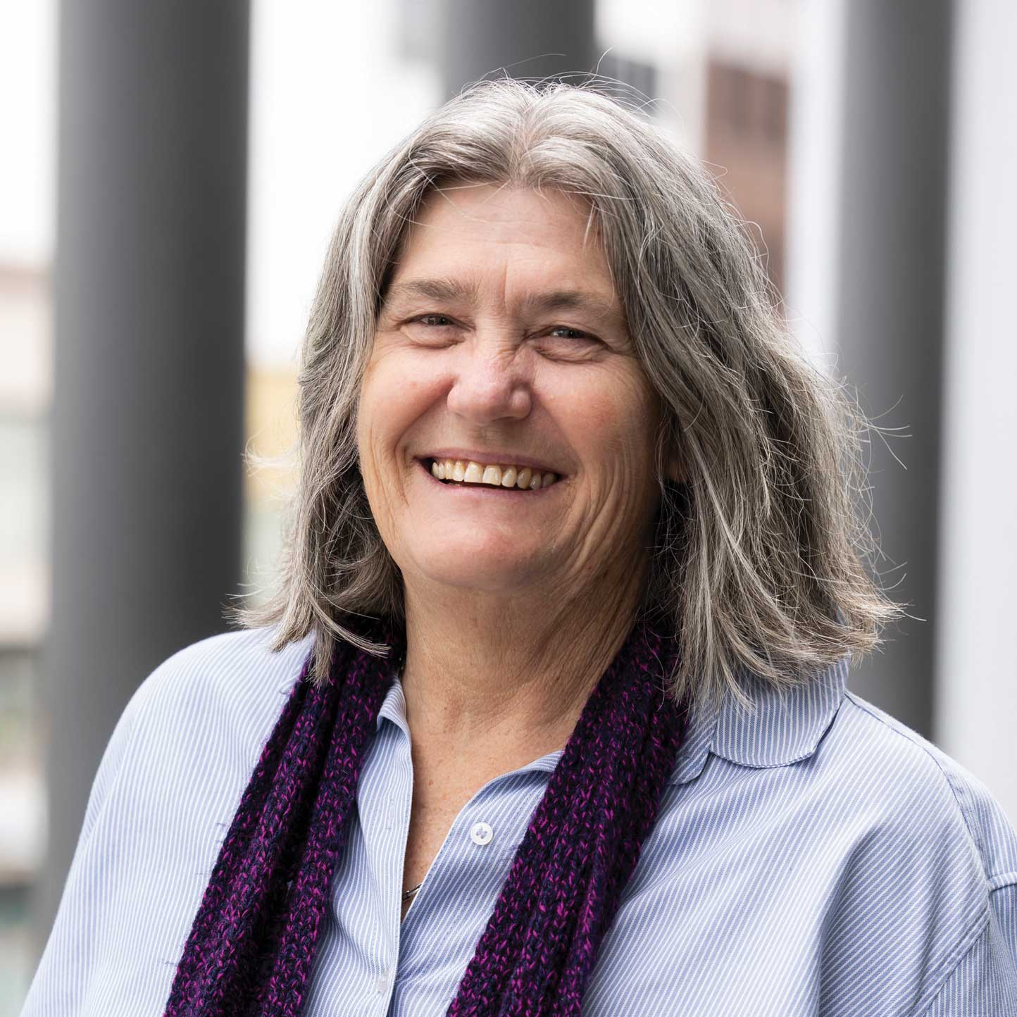 Head-and-shoulders portrait of a person with shoulder-length grey hair, wearing a light blue striped shirt and a dark knitted scarf. The photograph is taken outdoors, with blurred architectural columns and building features in the background.