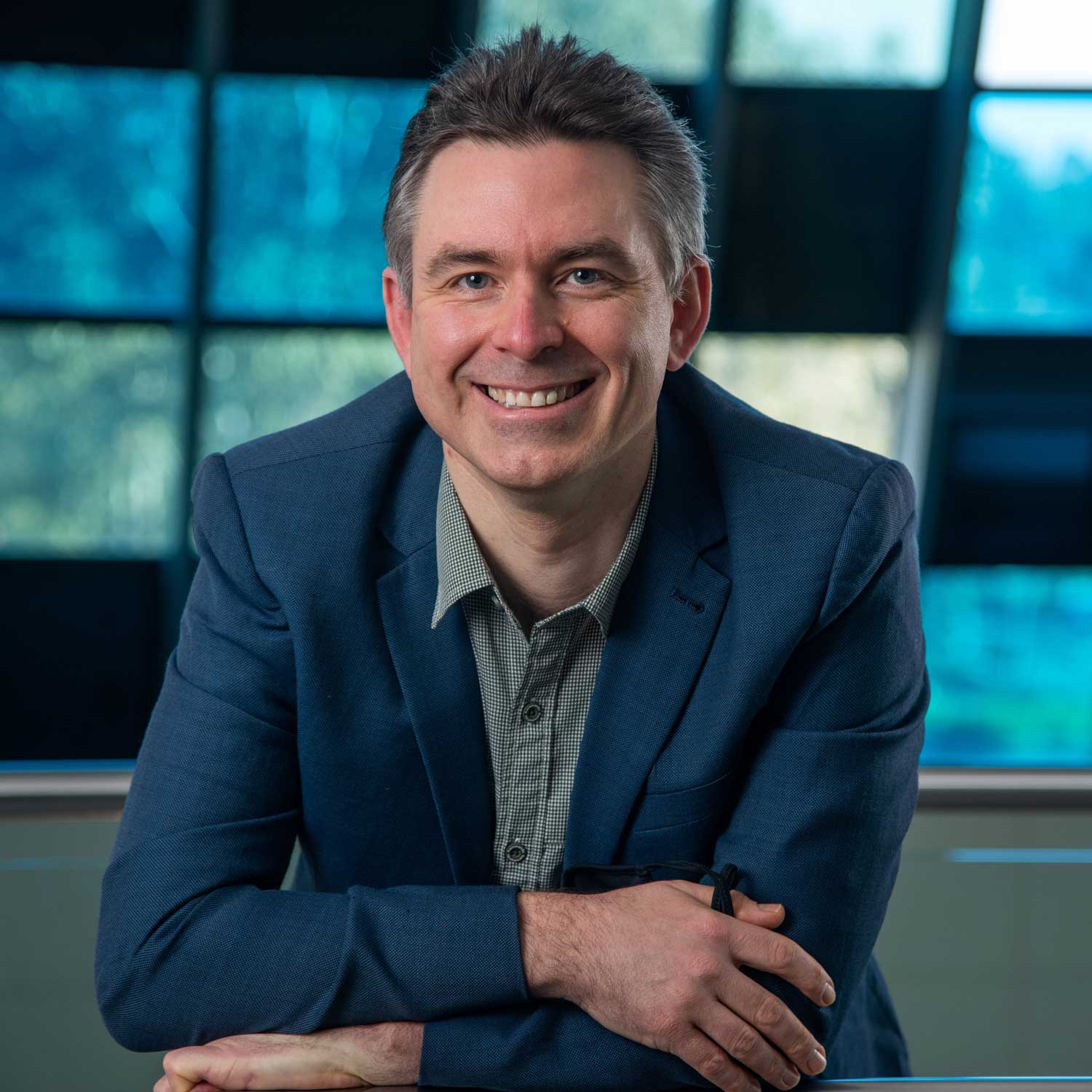 Professional head-and-shoulders portrait of Dr John Ward wearing a blue blazer and collared shirt, seated indoors with folded arms resting on a surface. Large blue-tinted windows in the background.