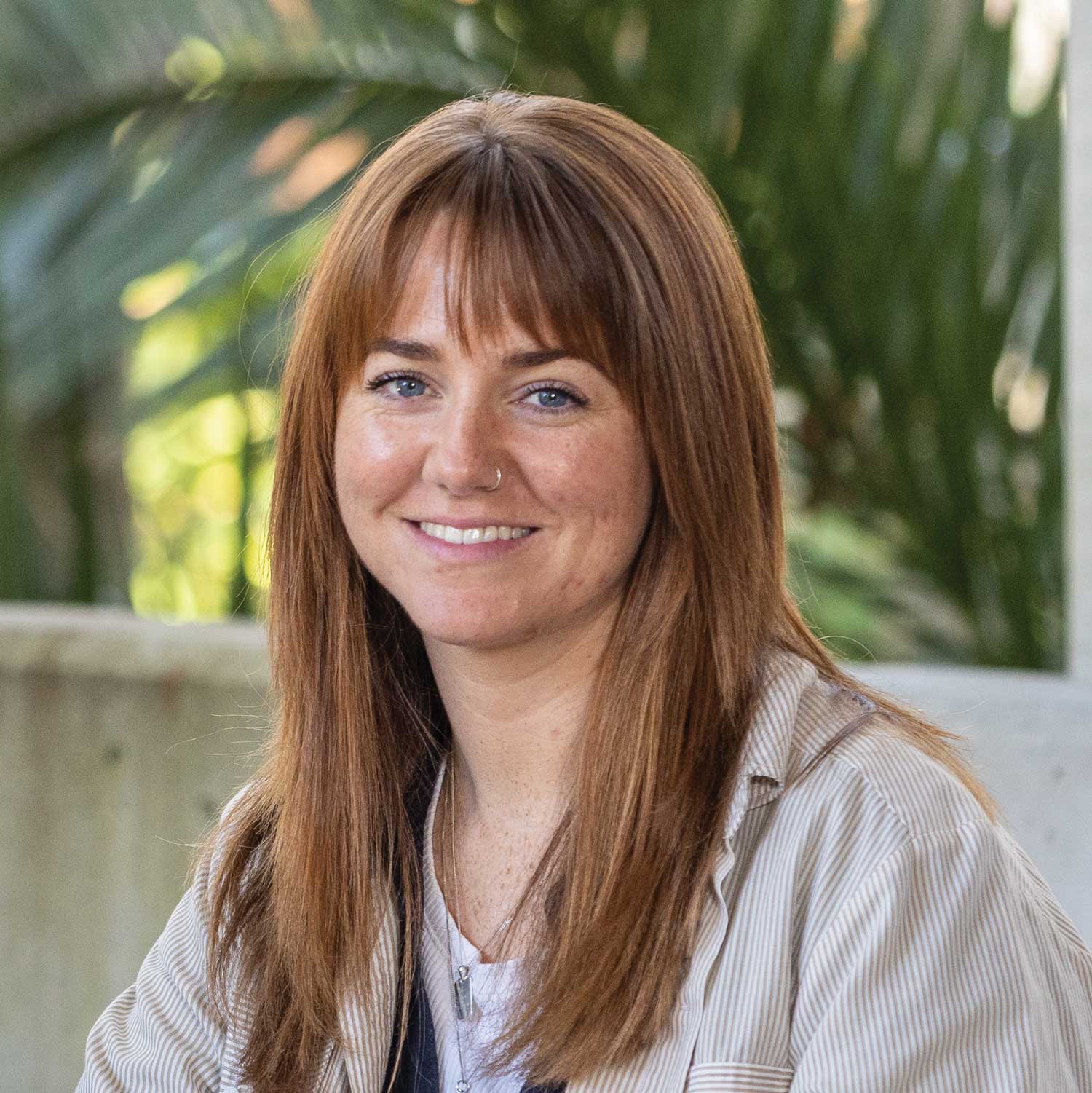 Professional head-and-shoulders portrait of Georgia Young with long copper hair, wearing a light button-up shirt over a white top. Georgia is seated outdoors or in a garden-like setting, with soft natural light and blurred green foliage in the background.