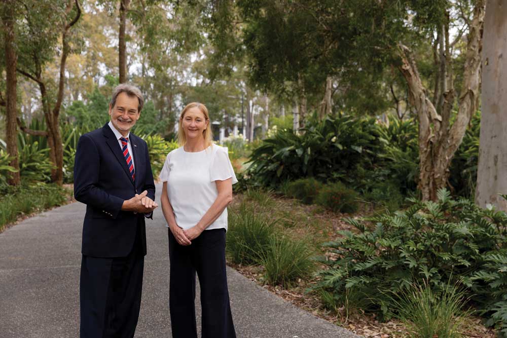 John Dugas and Julie Thomas-Bailey stand on a tree-lined pathway surrounded by native greenery and landscaped gardens, posing for a photograph in an outdoor campus setting.