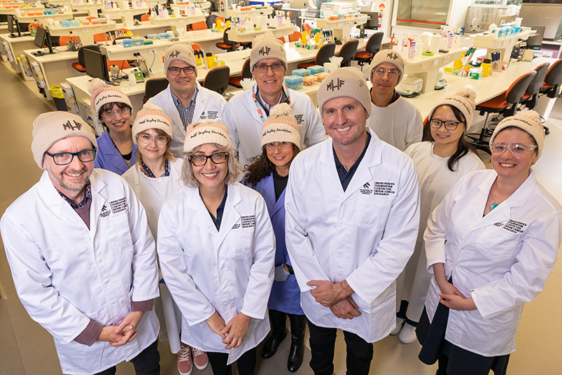 A group of people look up at the camera in a lab setting. They're wearing lab coats and Mark Hughes Foundation beanies