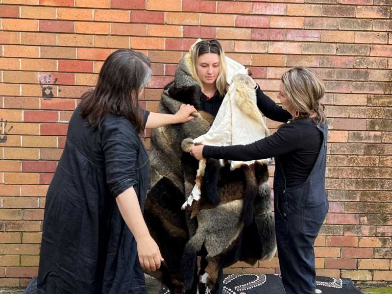 Two female adults wrapping a high school student in a long possum skin cloak
