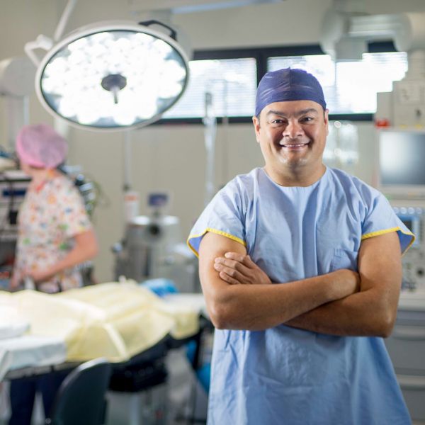 Dr Kelvin Kong is wearing scrubs and smiling with his arms crossed in a surgery room with nurses in the background. Celebrating our outstanding community with 2024 Australia Day Honours .