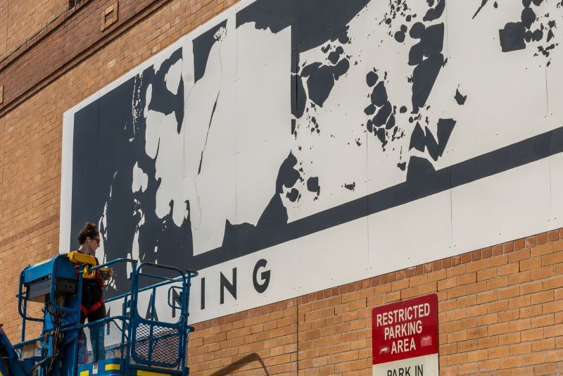 Large black and white mural on brick wall painted by artist on scissor lift