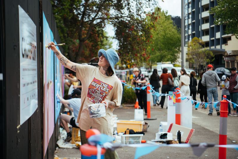 Woman painting a mural on a wall in front of a crowd on a busy street