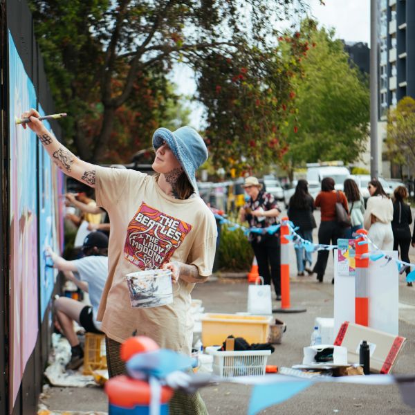 Woman painting a mural on a wall in front of a crowd on a busy street
