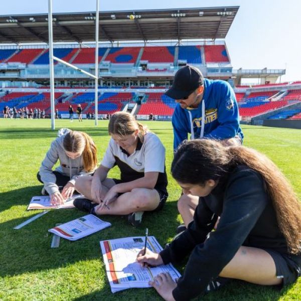 Aboriginal high school students sitting around with an undergraduate filling out numeracy booklet at MacDonald Jones Stadium as part of the Aboriginal Numeracy Gala Day. Students increase cultural understanding with numeracy and Knights