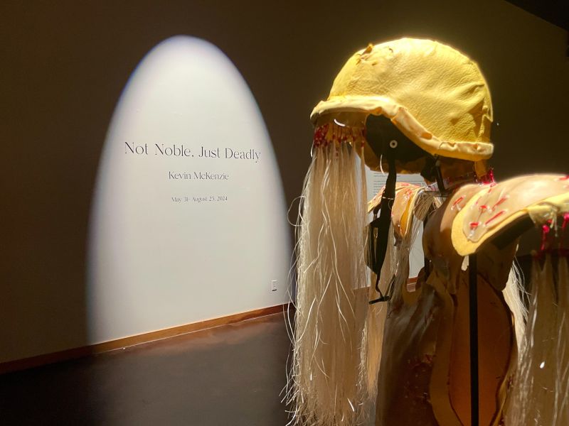 Yellow helmet and body art display with straw tassles hanging down from red twine in an art gallery. There is a white wall behind it that says "Not Noble. Just Deadly. Kevin McKenzie May 31 - August 25 2024."