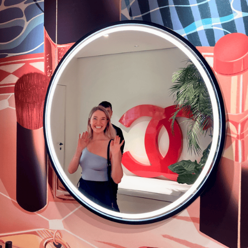 Izzy is smiling and waving in front of a large circular mirror with the Chanel logo in the background. She is surrounded by various beauty products and her designs on display.