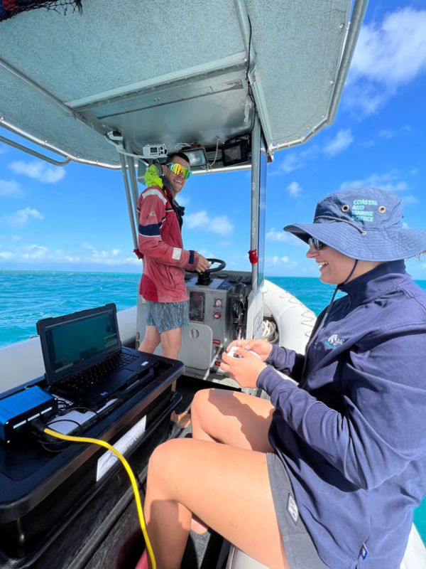 Two researchers on a boat with equipment, one operating the boat and the other, Kristen, piloting an underwater drone, in a sunny setting with clear blue skies and ocean.