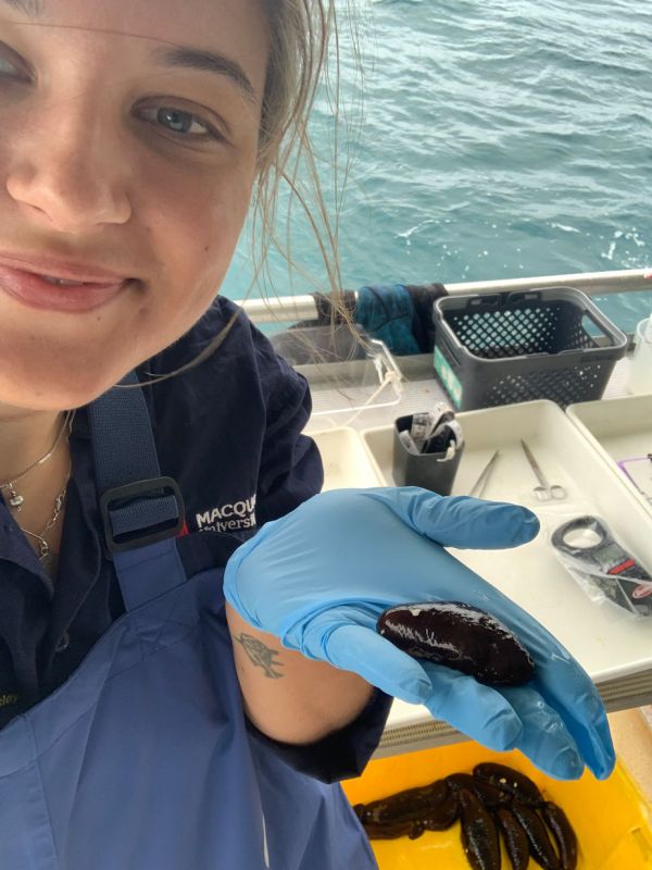 Kristen holds a sea cucumber on a boat, with marine research equipment visible in the background.