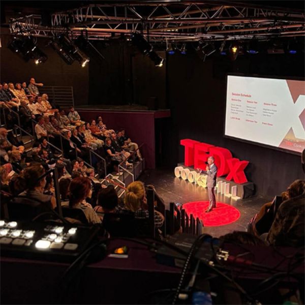 A speaker stands on stage at a TEDx event, addressing an audience with the TEDx logo and session schedule projected on a screen behind them.