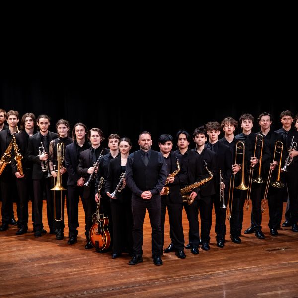 A large group of musicians in black attire holding brass and woodwind instruments stands on a wooden stage with a conductor at the center.