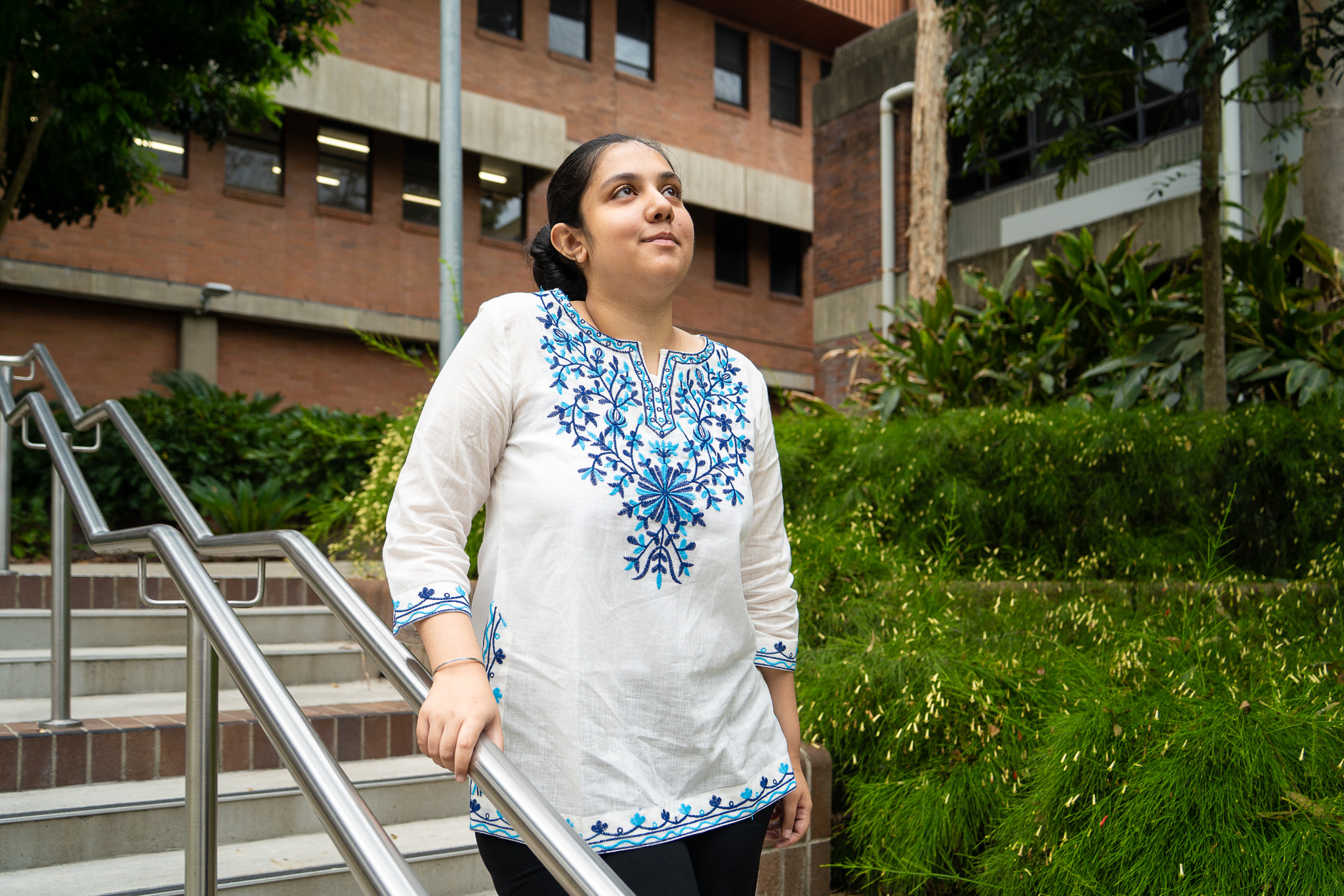 A person stands outdoors on a stairway railing in front of a brick university building, wearing a white three‑quarter‑sleeve tunic with intricate blue floral embroidery across the chest, sleeves, and hem. Lush green plants line the pathway beside them, and windows and metal railings are visible in the background, creating a campus setting.