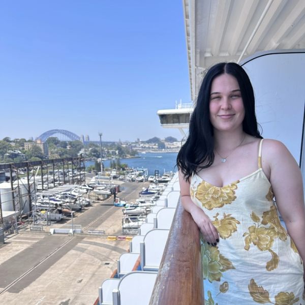 A woman with long dark hair stands on a cruise ship balcony, wearing a floral dress. She smiles, with a clear blue sky and distant cityscape in the background.