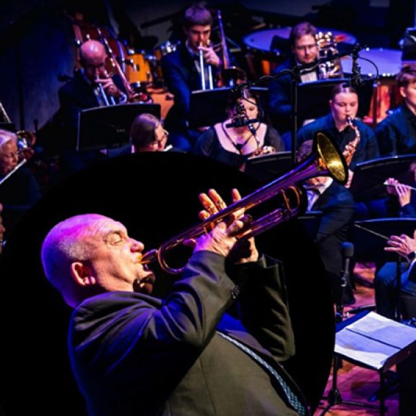 A musician in the foreground plays a trumpet on stage while a large seated ensemble of wind and brass instrumentalists performs behind.