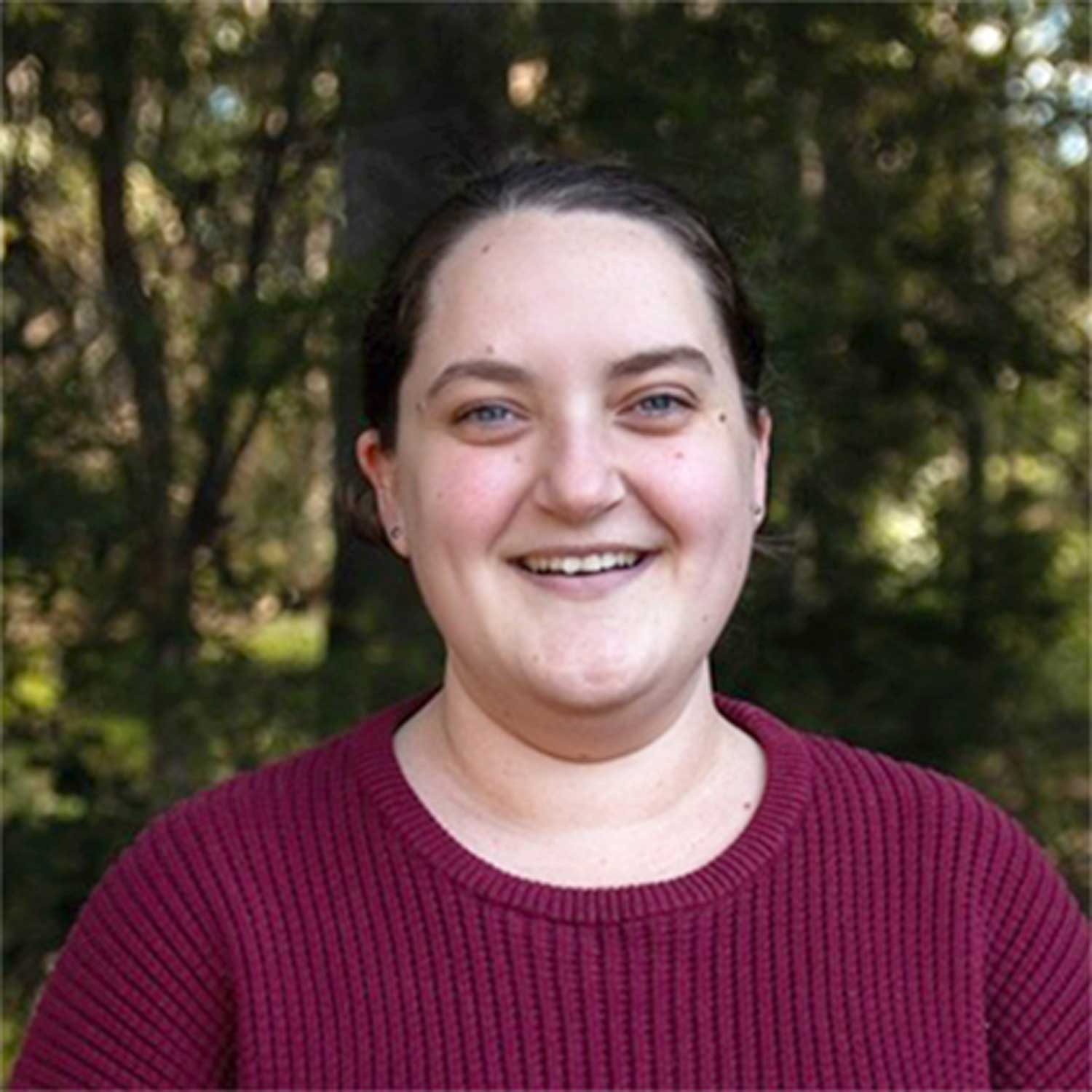 Brunette female smiling at the camera, pictured outdoors with greenery in the background.