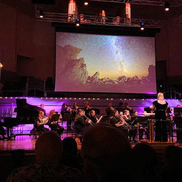 An orchestra performs on a concert hall stage with a conductor at the front, while a large screen behind the musicians displays a star-filled night sky over rocky landscapes.
