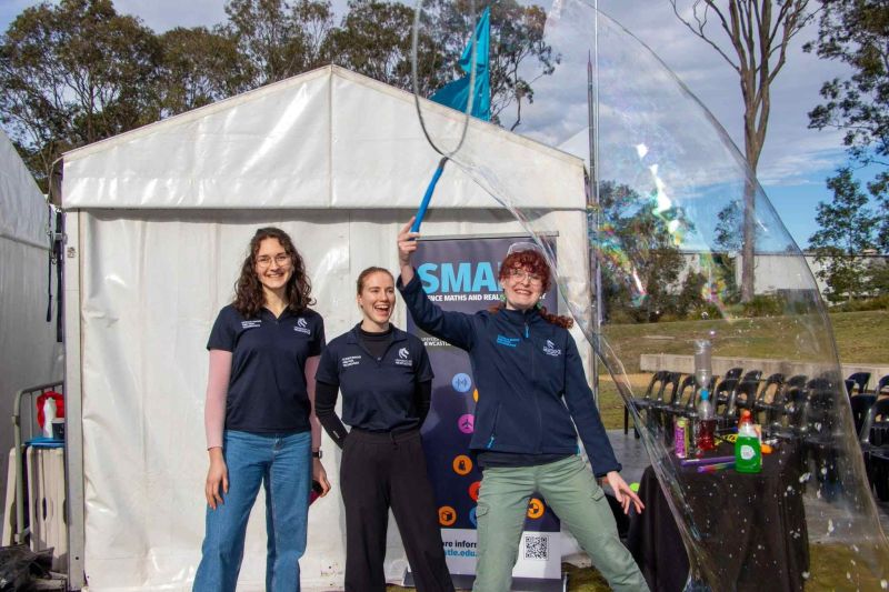 3 women in front of a white tent. 1 women with giant bubble 
