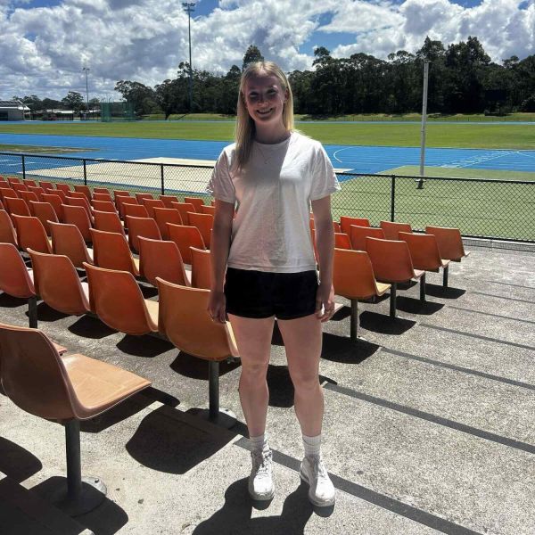 Woman with blonde hair standing in the seated section of a sports stadium smiling.