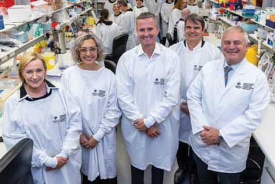 Five people wearing white laboratory coats stand together in a research laboratory. Laboratory benches, scientific equipment and researchers at work are visible in the background, highlighting an active medical research environment.