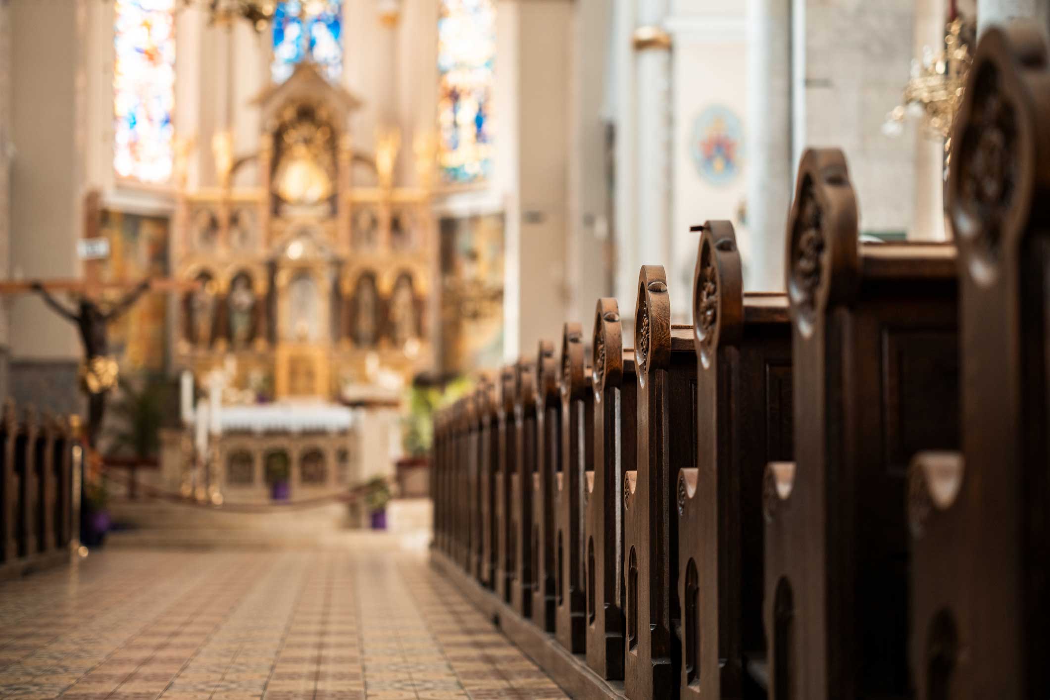 Interior view of a church with rows of dark wooden pews lining a central aisle. The aisle leads toward an ornate altar and stained-glass windows, softly blurred in the background, with patterned floor tiles and warm natural light throughout the space.