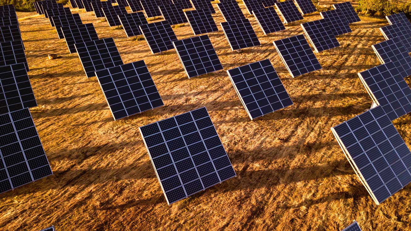 Aerial view of a large solar farm featuring rows of dark solar panels installed across a dry, golden-brown landscape. The panels are arranged in a grid pattern and cast long shadows in warm sunlight, highlighting a large-scale renewable energy installation.