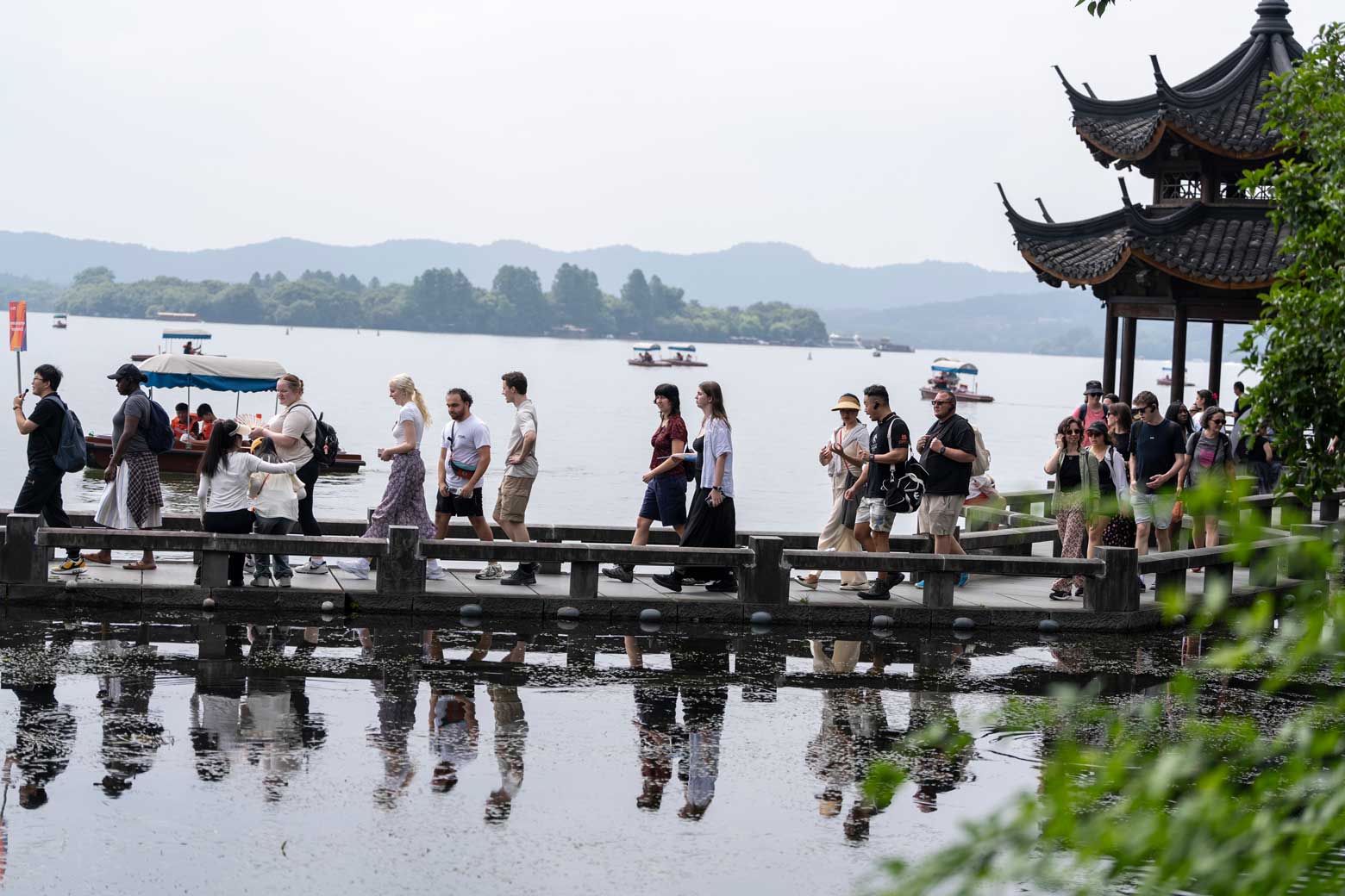 A group of people walk along a waterside boardwalk beside a traditional Chinese-style pavilion. The boardwalk extends over a reflective pond, with a large lake, small boats, tree-covered islands and distant hills visible in the background.