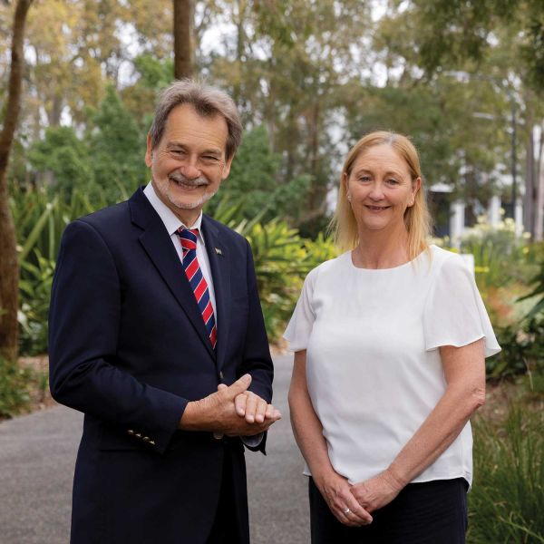 John Dugas and Julie Thomas-Bailey stand on a tree-lined pathway surrounded by native greenery and landscaped gardens, posing for a photograph in an outdoor campus setting.. A lifetime of giving back