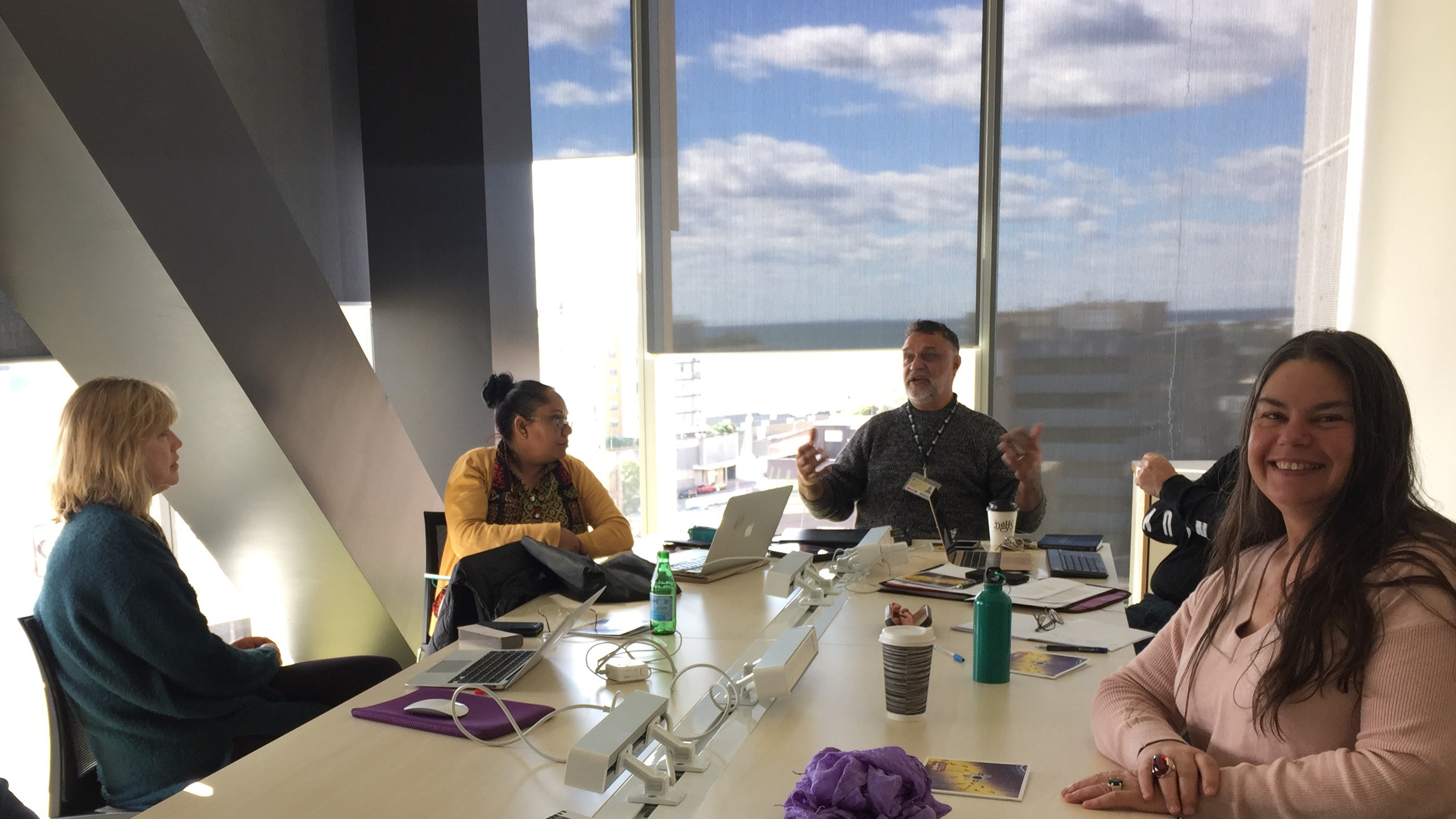 People sitting around a table. Woman smiling at camera. Man at end speaking and gesturing with his hands. Three other people listening