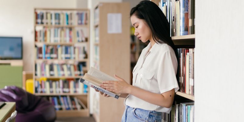 Woman standing against a bookshelf reading a book