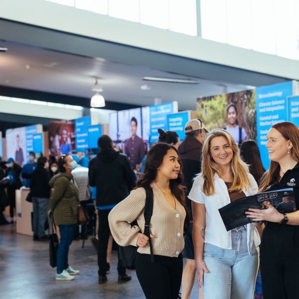 One woman holds an open day program to show two people. Open day stalls are visible in the background. People from across the region expected to get a taste of university life at Central Coast Open Day
