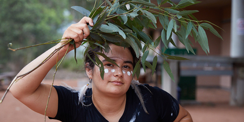 An Aboriginal person dances with some eucalyptus leaves, they have white dots painted across their cheeks 