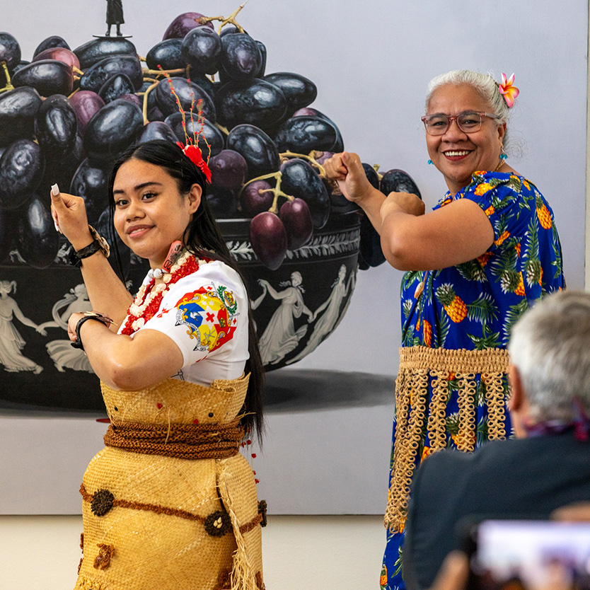 Two women dance wearing traditional Pacific dress in front of an artowkr of a bowl of grapes^empty:{ds__assetid^as_asset:asset_name}