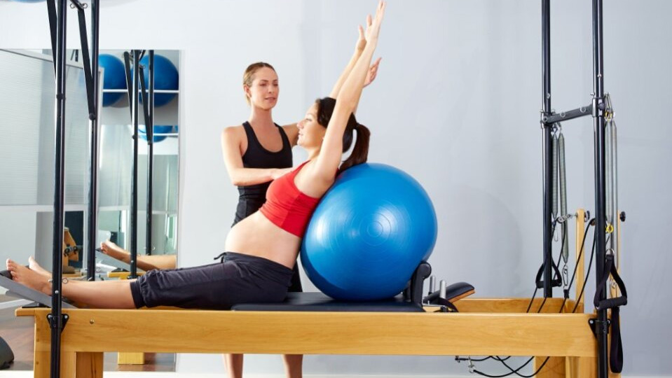 An instructor assists a pregnant women during a reformer pilates fitball exercise